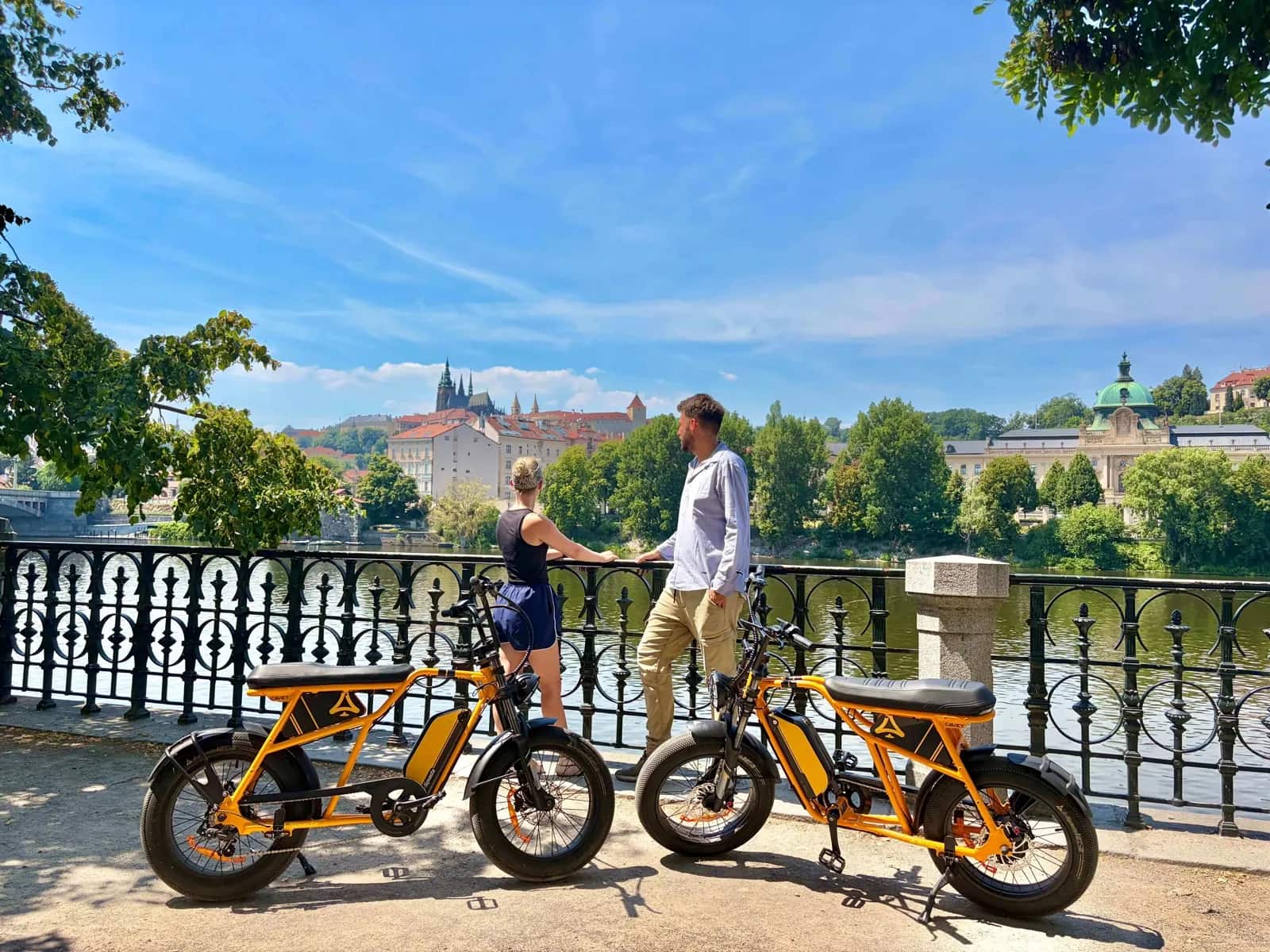 A couple with chopper e-bikes on the embankment, Prague Castle across the river