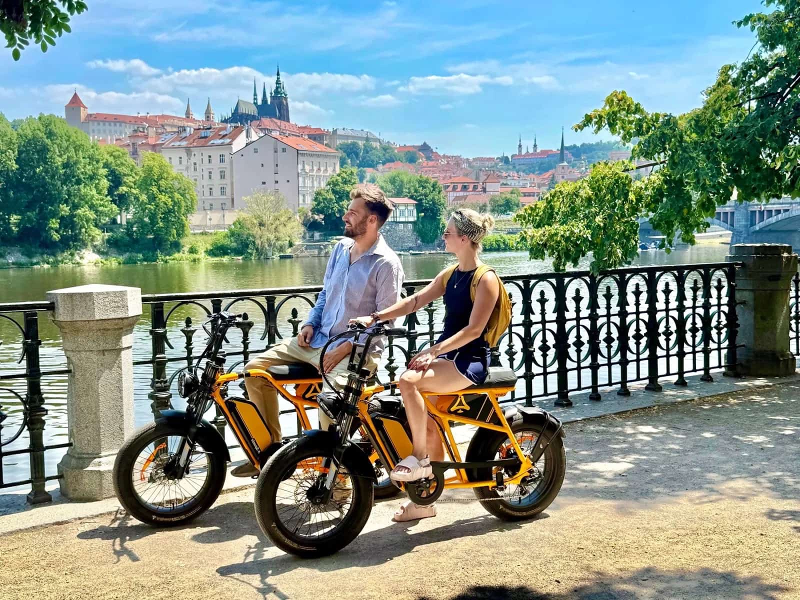 Two riders on orange chopper e-bikes by the river with Prague Castle behind